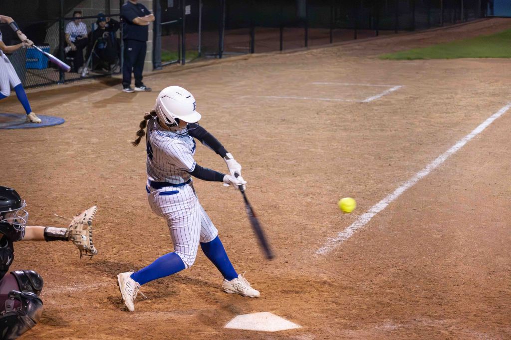A female softball player in a striped uniform is swinging a bat at a pitched ball on a dirt field, with a catcher in the foreground preparing to catch. The setting is illuminated for a night game.