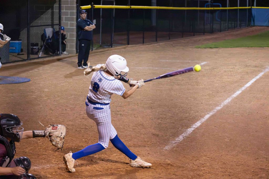 A young female softball player with long hair swings a bat at a yellow softball during a night game, with teammates and a coach visible in the background.