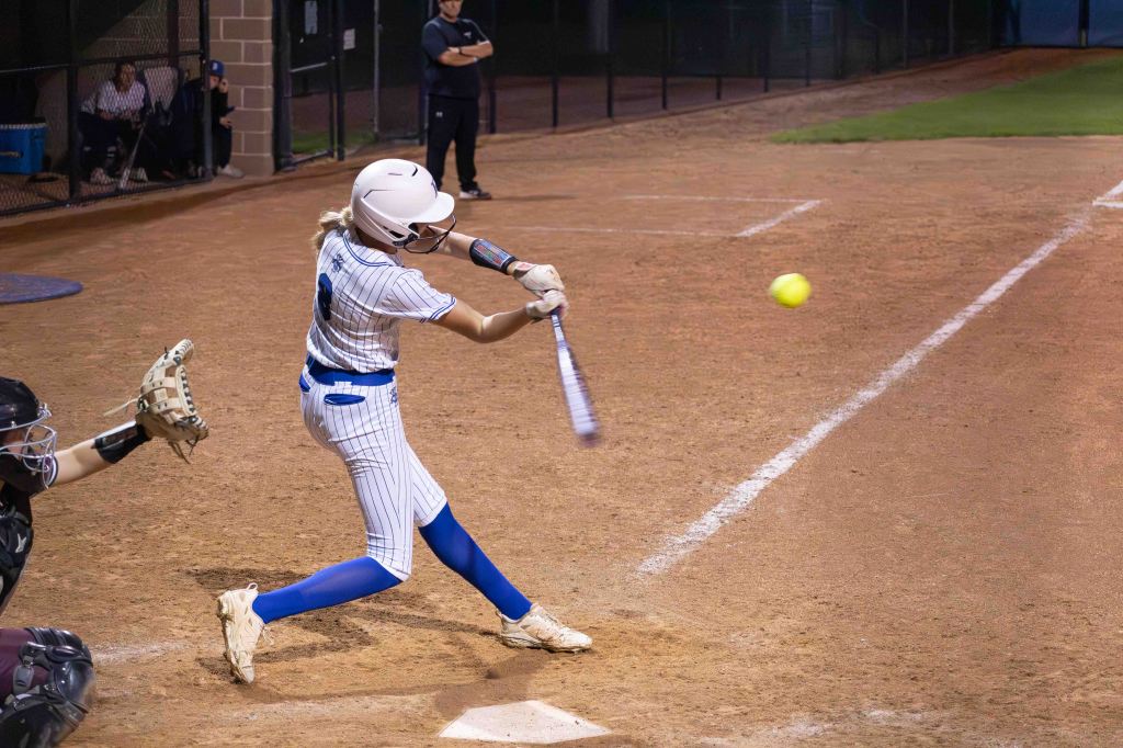 A female softball player in a striped uniform swings a bat at a flying softball in a night game, with a catcher in the foreground.