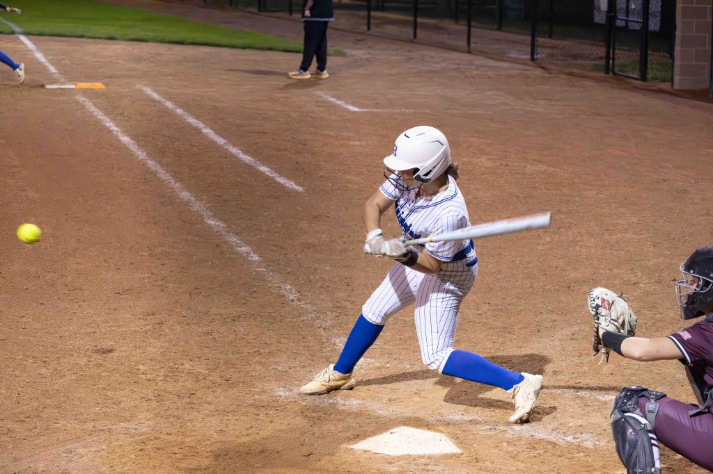 A softball player swinging a bat to hit a pitch during a game, with a catcher in position behind home plate.