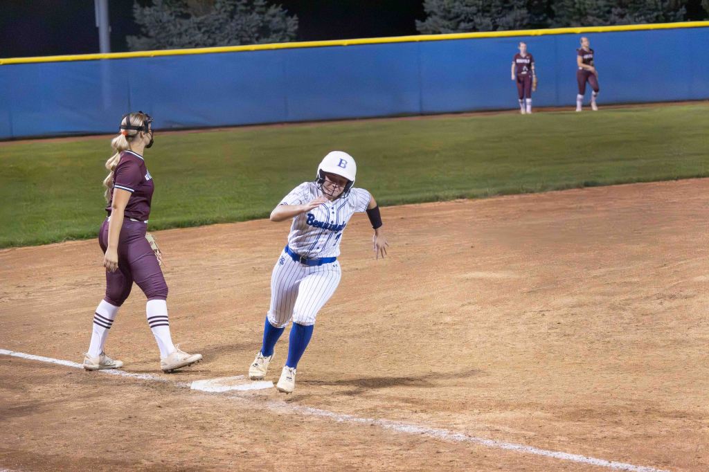A softball player in a blue and white striped uniform runs to first base while a player in a maroon uniform stands nearby. Nighttime setting with field markings visible.