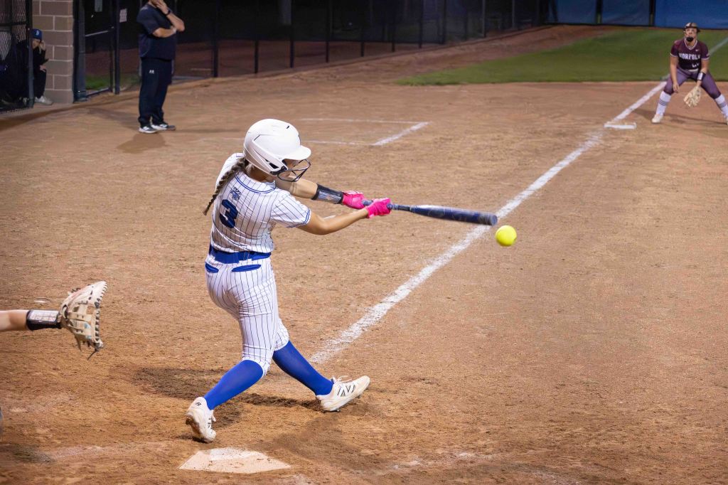 A young female softball player swinging a bat at a yellow softball during a game, with teammates and opponents in the background.