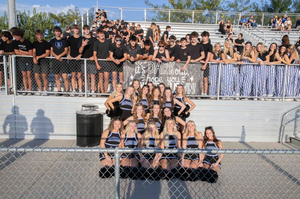 A group of cheerleaders in black and blue uniforms posing in front of a crowd of students in black shirts at a sporting event, with a banner that reads 'it's DARK out, hope you brought your SUN!'