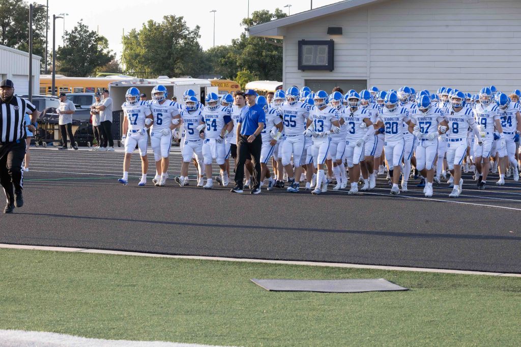 A football team in white and blue uniforms marching onto the field, led by a coach and referee, with spectators observing in the background.