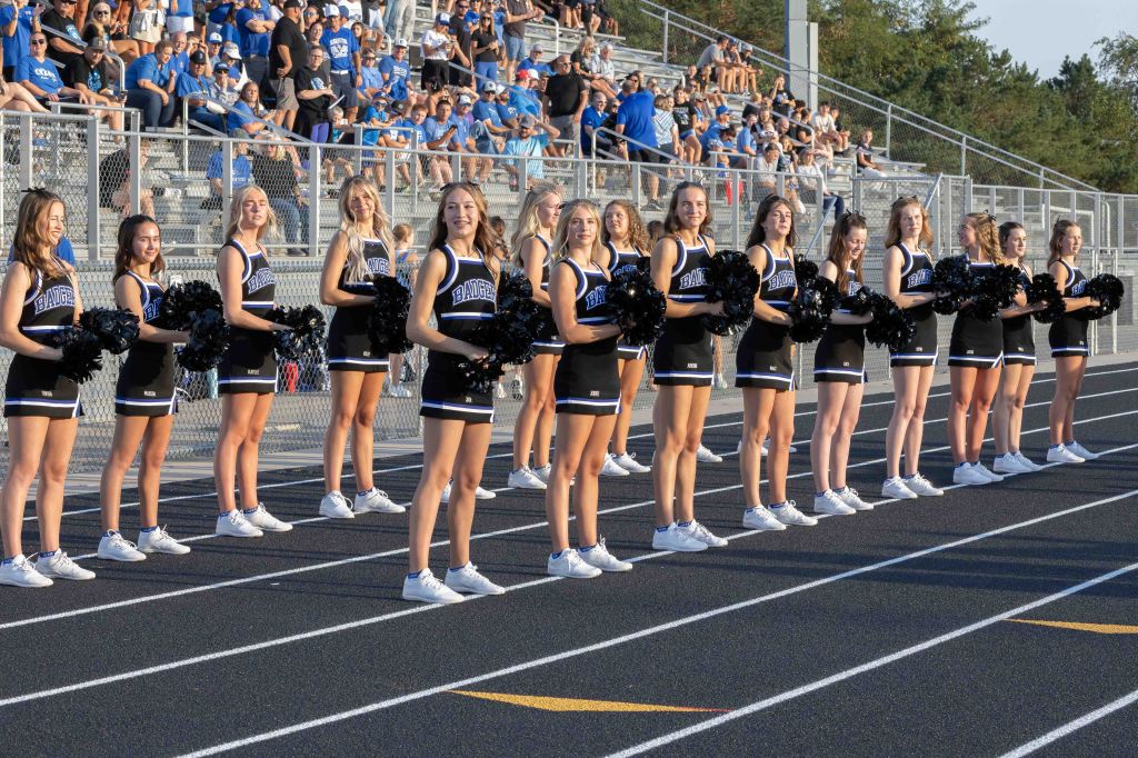 A cheerleading squad in matching uniforms stands on the sidelines of a sports field, performing with pom-poms for an audience in the background.