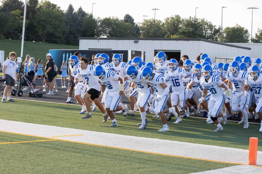 A football team in blue and white uniforms rushes onto the field, with players displaying enthusiasm and energy as they prepare for a game.