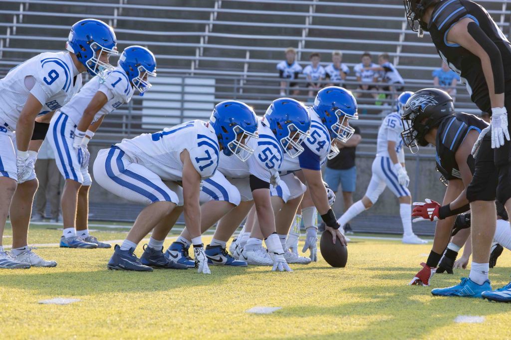 A youth football game featuring players in blue and white uniforms preparing for a play, with an opposing team in black uniforms facing them.
