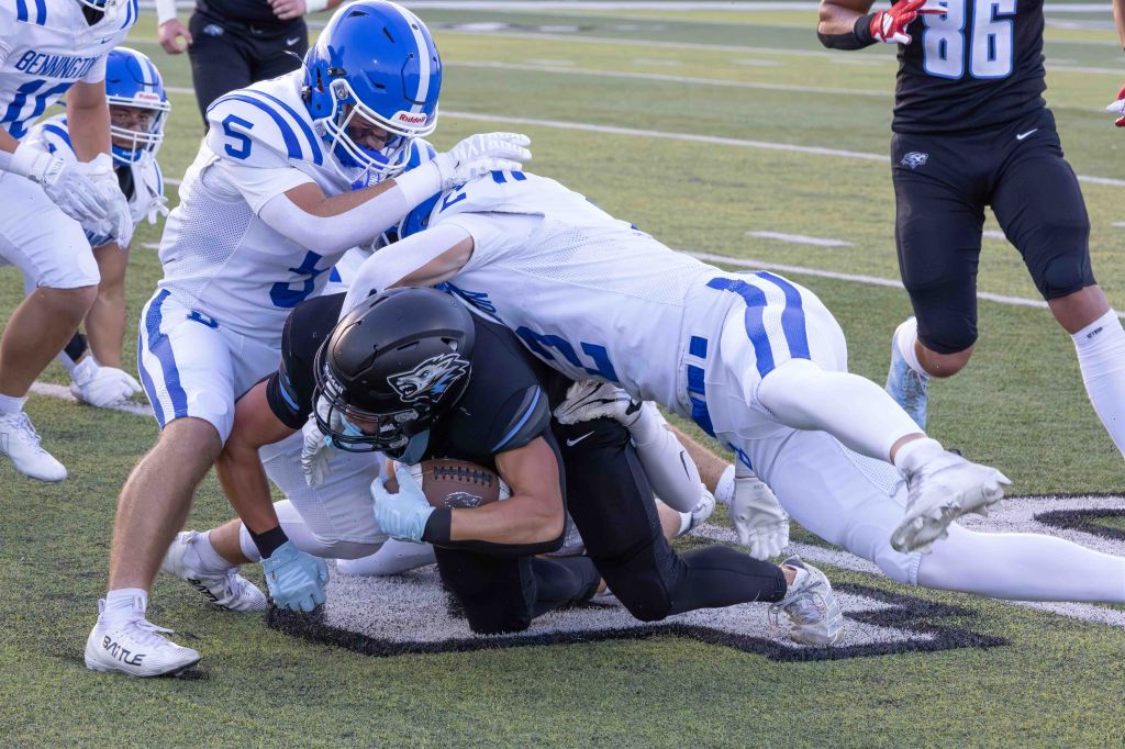 Two football players in blue uniforms tackle a player in a black uniform on the field, with additional players in the background.