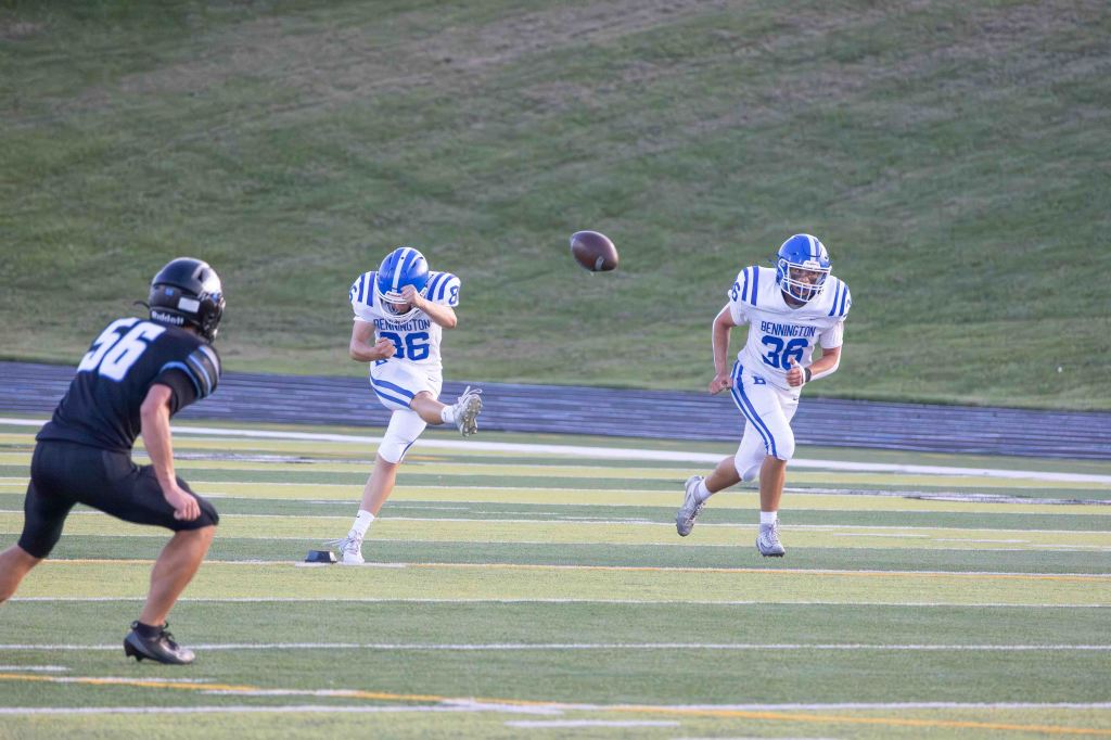 A high school football player in a white and blue uniform kicks a football while another player runs behind him. An opposing player in black is positioned to the left, observing the play.
