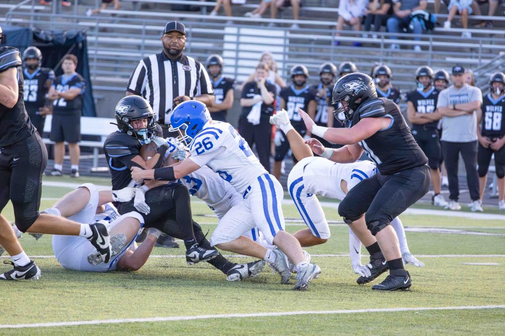 Action shot of a high school football game with players in black and blue uniforms tackling each other on the field.