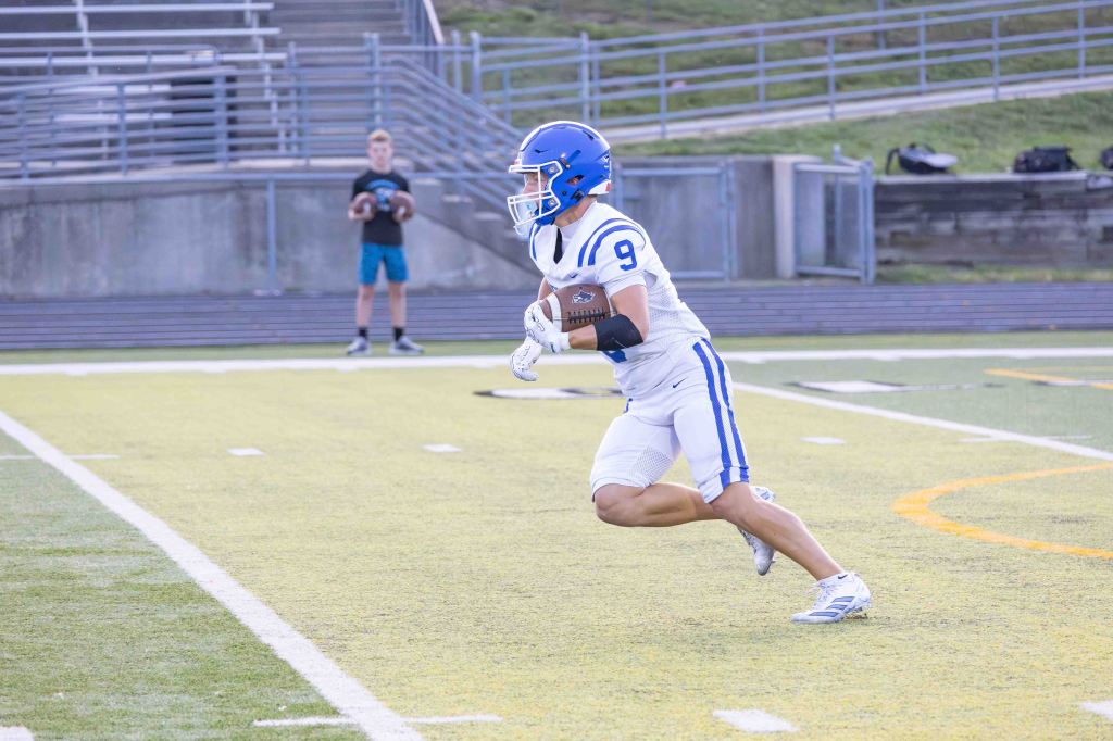 A football player in a blue helmet and white uniform runs on the field, holding a football, with another player in the background.