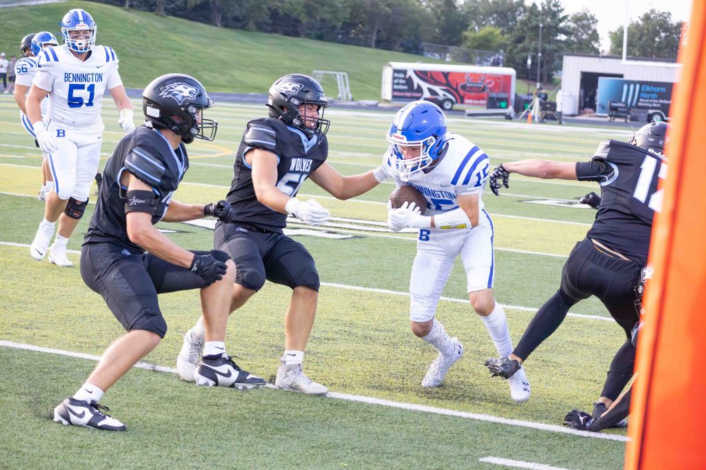 A high school football game in progress, featuring players in blue and white jerseys from Bennington and black jerseys from the opposing team. A Bennington player is being tackled while another teammate runs in the background.