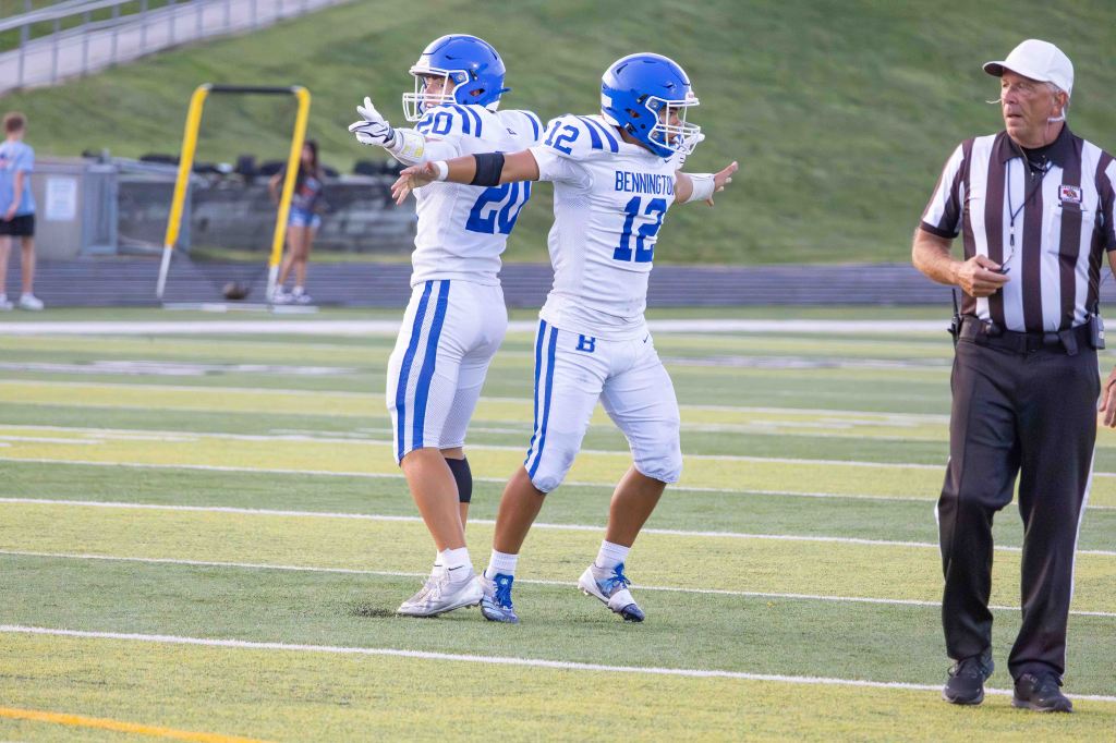 Two football players in white uniforms with blue accents celebrating on a field, with a referee observing nearby.