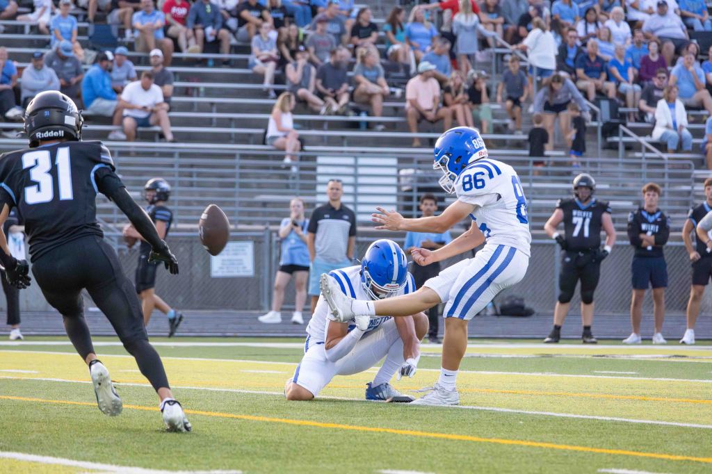 A football player wearing blue and white uniform prepares to kick a football that is being held by a teammate on the field, with spectators in the background.