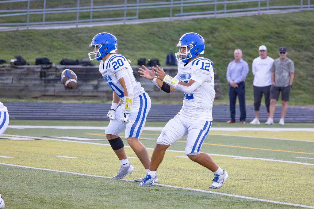 A quarterback in a white football uniform with blue accents prepares to throw a football, while a teammate in a similar uniform runs nearby.