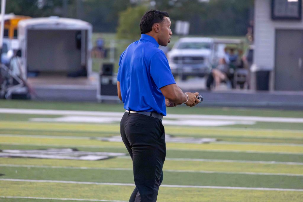 A coach in a blue shirt and black pants stands on a football field, holding a device and looking towards the game.