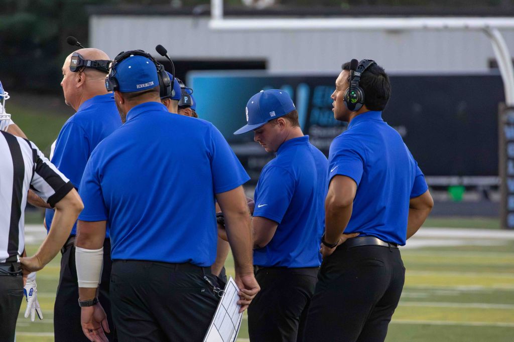 Group of football coaches in blue uniforms discussing strategy, with a referee visible in the foreground.