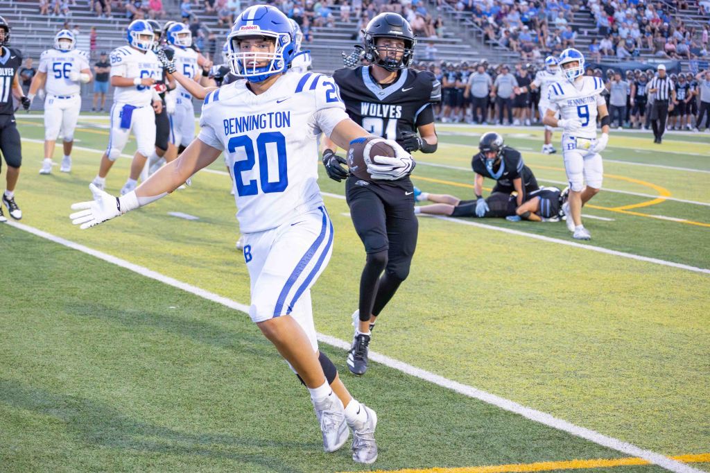A football player from Bennington, wearing jersey number 20, celebrates after a play on the field. In the background, players from both teams are visible, with the opposing team in black uniforms and spectators in the stands.