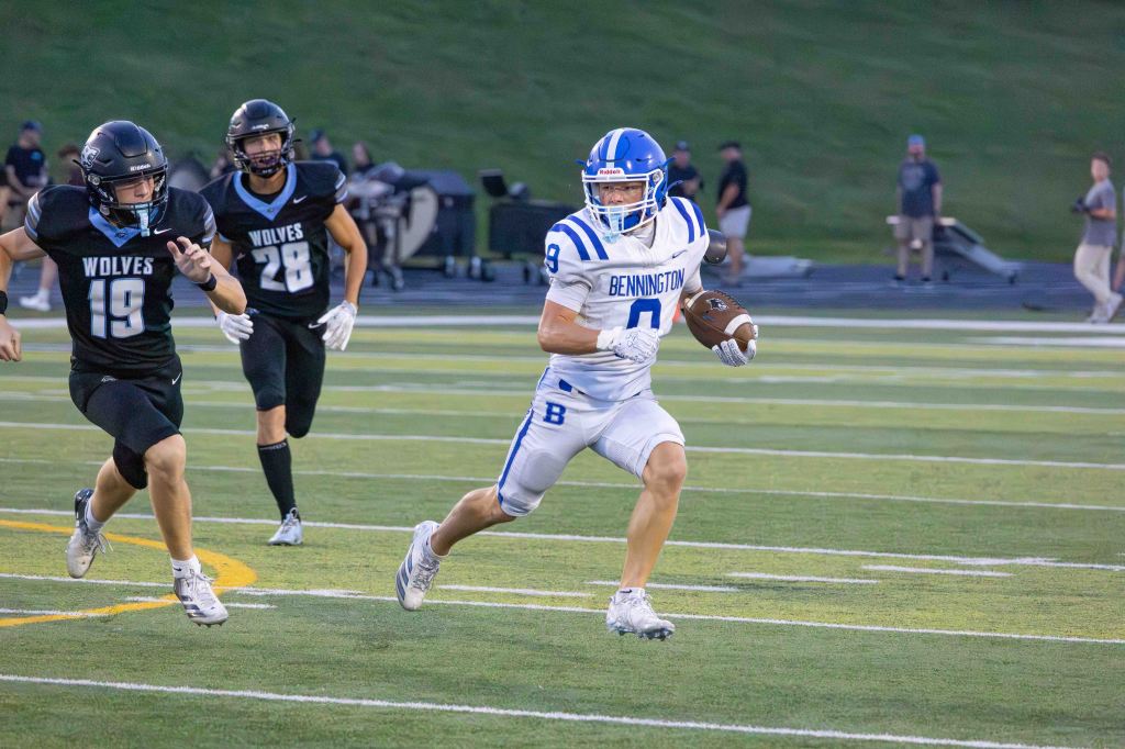 A high school football player in a blue and white uniform labeled 'BENNINGTON' runs with a football while being chased by two players in black uniforms labeled 'WOLVES' during a game.