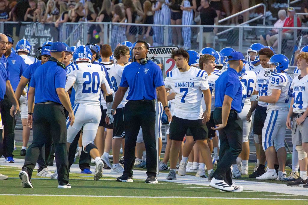 A group of high school football players in blue and white uniforms are on the sidelines during a game. Coaches and team staff are engaged in discussions as players prepare for their next play.