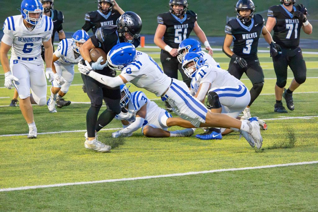 A football player in blue and white uniform attempts to tackle an opposing player in black during a game, with other players in the background.