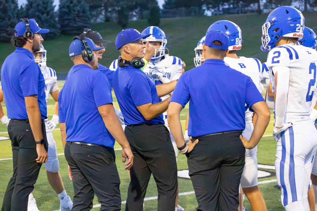 A group of football coaches wearing blue shirts gather around players in white uniforms, discussing strategies on the field.