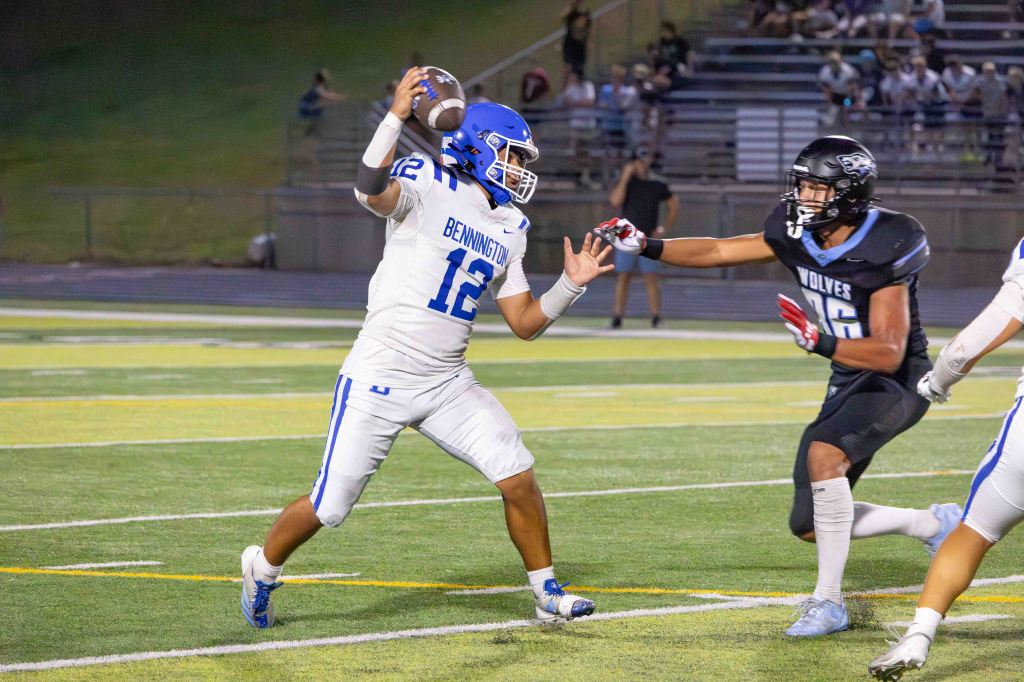 A football player in a white jersey with blue accents is attempting to throw a football while being pursued by a defender in a black jersey. The scene takes place on a football field during a game, with spectators in the background.