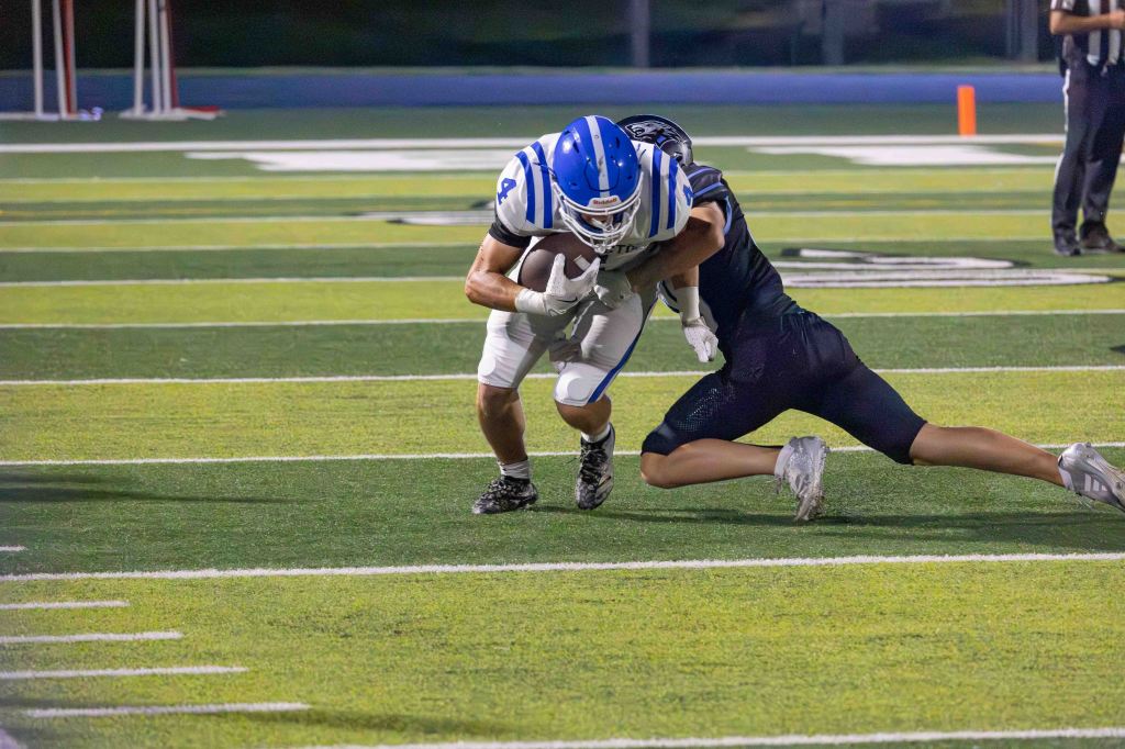 A football player in a blue and white uniform is being tackled by an opponent wearing a black uniform on a green field.