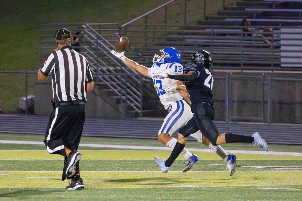 A high school football player in a blue uniform attempts to catch a ball while being defended by a player in a black uniform. The referee observes the play in the foreground.