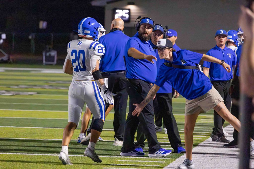 A football player wearing jersey number 20, walks along the sideline as coaches and staff wearing blue shirts engage with him during a game.
