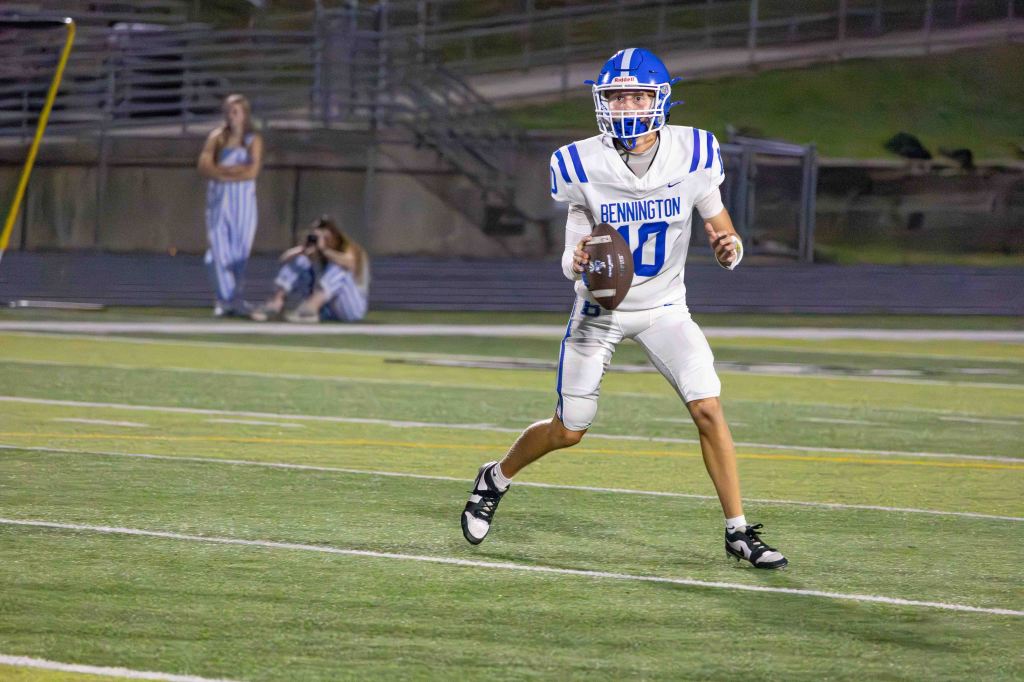 A quarterback in a white football uniform with blue stripes, wearing a helmet, is running on a field while holding a football. In the background, a few people are watching the game.