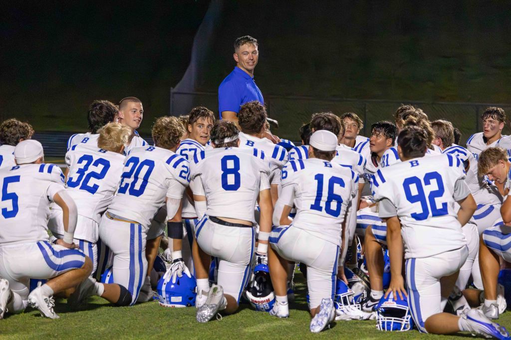 A high school football team in white uniforms with blue stripes gathers around their coach for a team talk after practice.