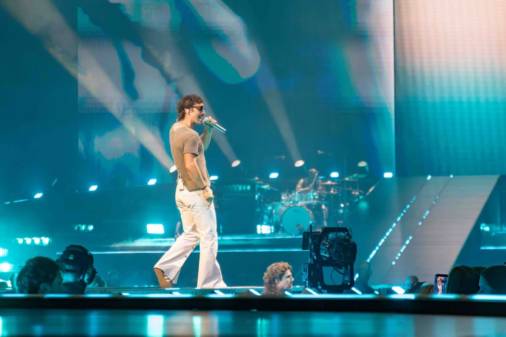 A performer singing on stage with a microphone, wearing sunglasses and a gray shirt, in front of a colorful light display, with a drummer visible in the background.