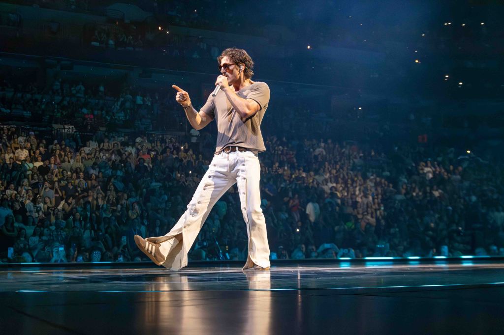 A male performer on stage, wearing a gray shirt and white pants, engaging with the audience during a concert, with a large crowd visible in the background.