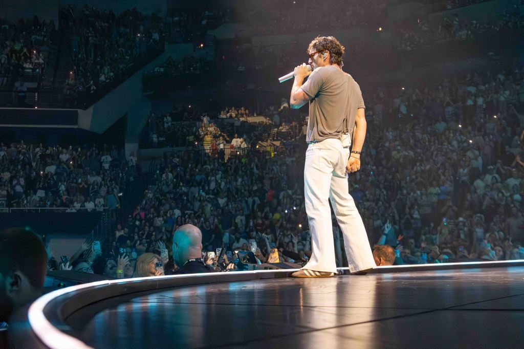 A performer sings on stage in front of a large crowd at a concert, with audience members holding up their phones to capture the moment.