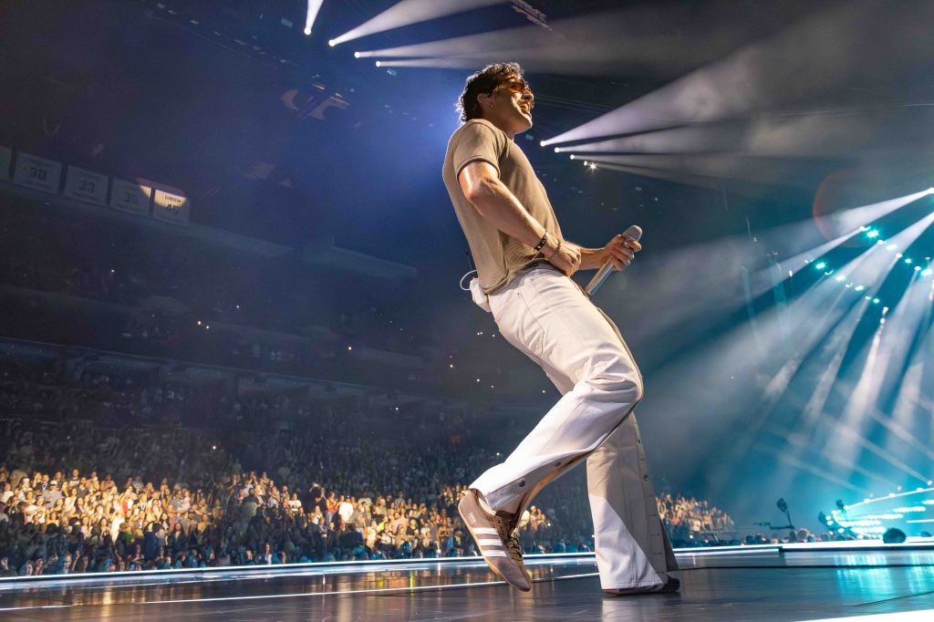 A male performer dances on stage while holding a microphone, illuminated by bright stage lights, with a large audience visible in the background.