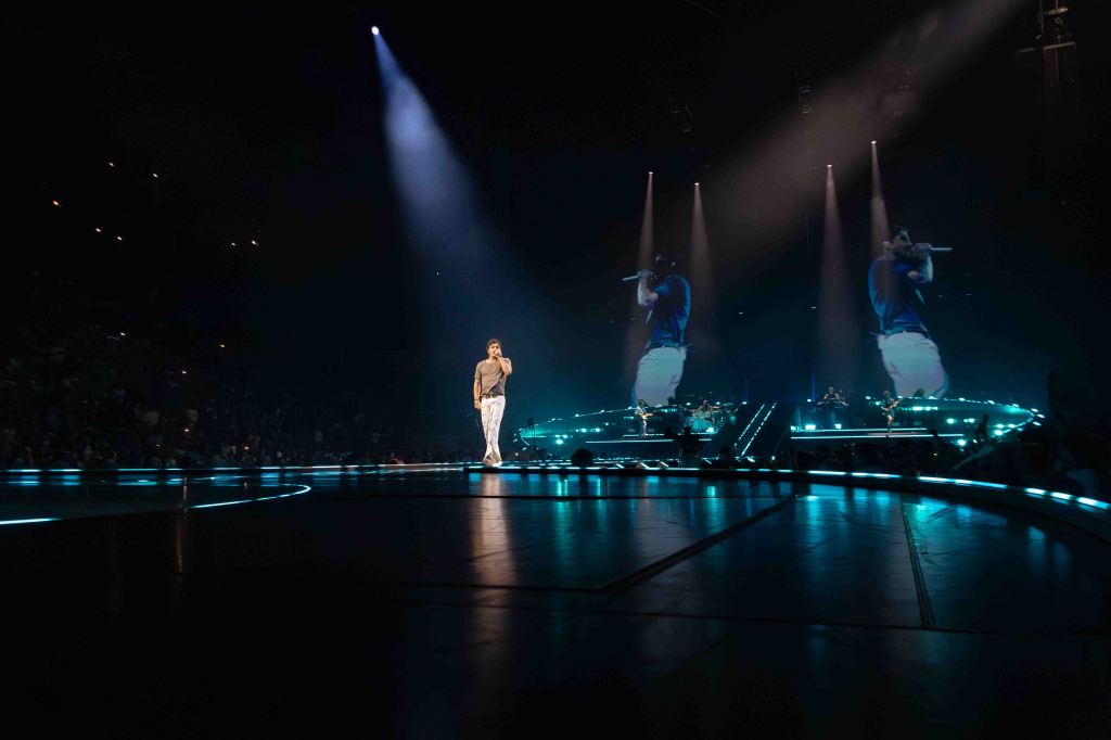 A performer on stage during a concert, illuminated by stage lights with large screens displaying their image in the background and an audience visible in the dimly lit venue.