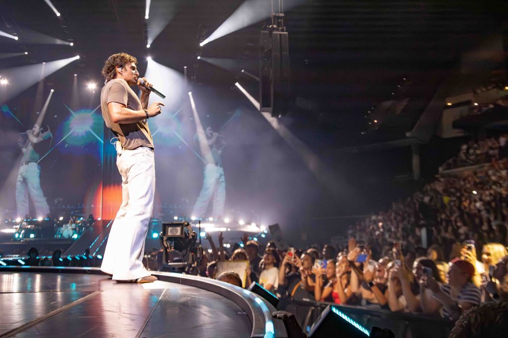 A male performer singing into a microphone on stage, wearing a gray shirt and white pants, with a large crowd of fans holding phones in the foreground. Bright stage lights and a colorful background enhance the concert atmosphere.