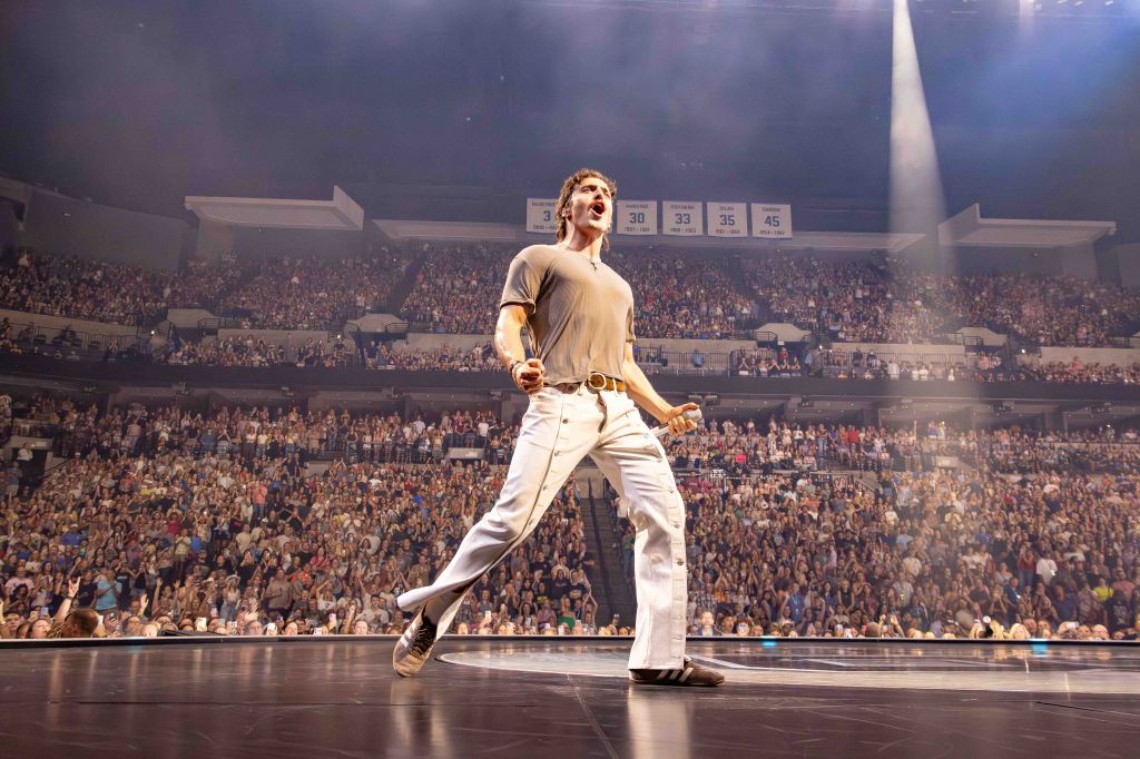 A performer energetically engages with a large crowd during a live concert, standing on stage with a microphone and dramatic lighting.