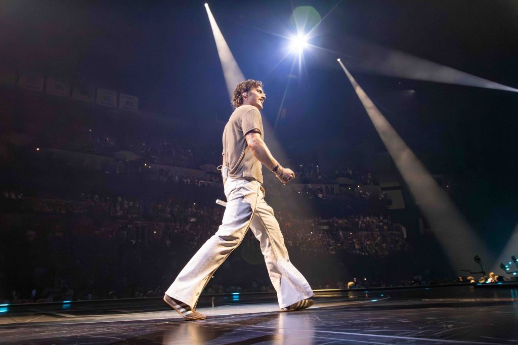 A performer walking across the stage during a concert, with bright stage lights and a cheering crowd in the background.