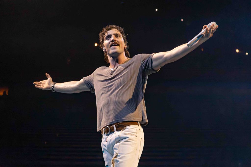 A performer with hands outstretched, wearing a gray shirt and white pants, singing passionately on stage with dim lighting and an audience visible in the background.