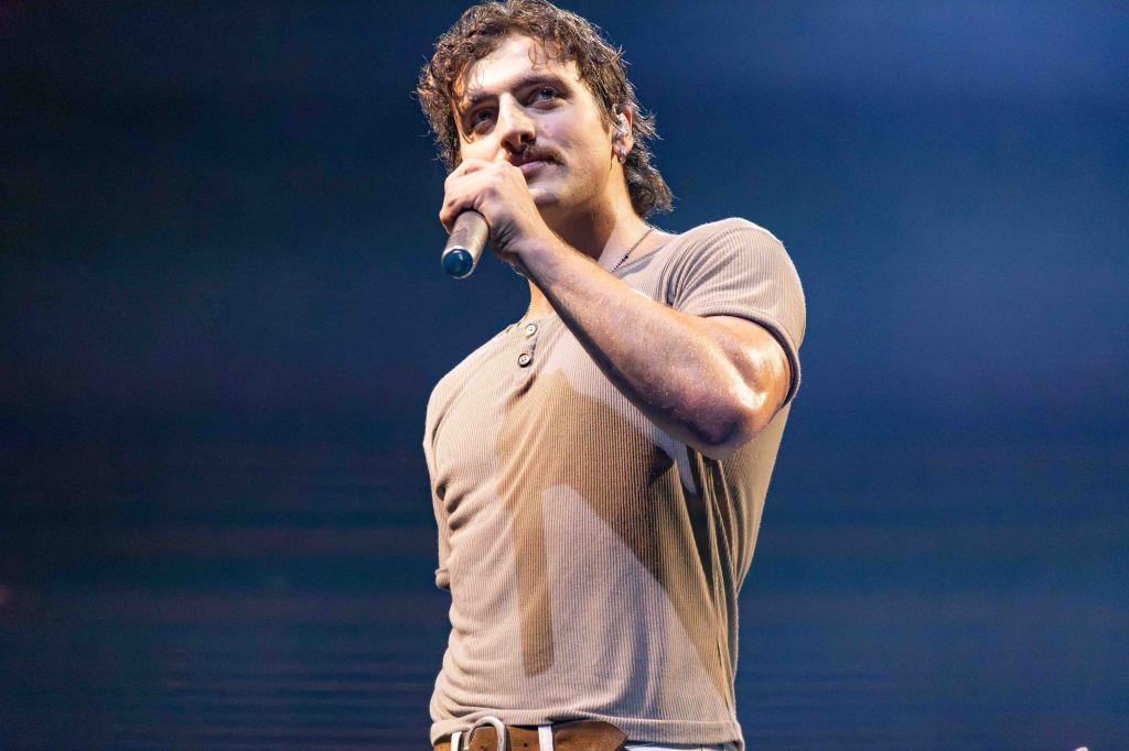 A male singer performing on stage with a microphone, wearing a fitted shirt and showing an intense expression.