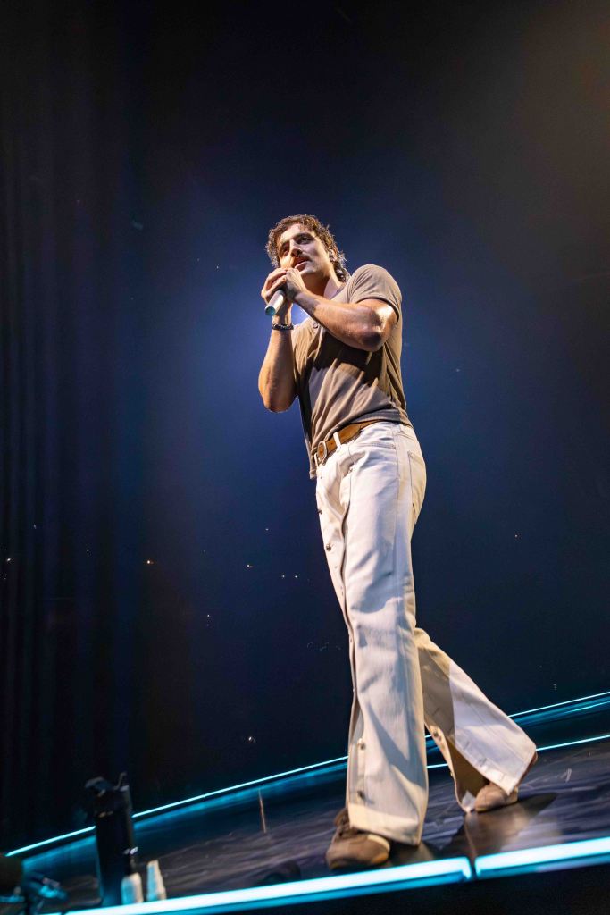 A performer on stage holding a microphone, wearing a fitted t-shirt and wide-leg pants, with stage lights creating a dramatic atmosphere.