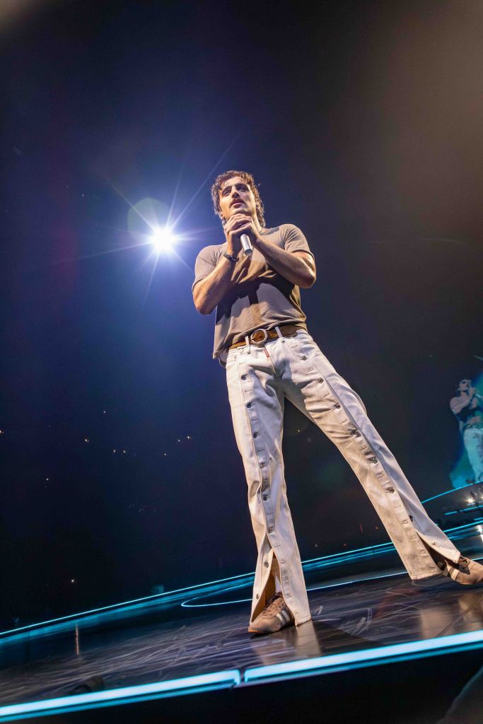 A performer on stage, wearing a fitted t-shirt and wide-legged pants, holding a microphone with a spotlight shining down.