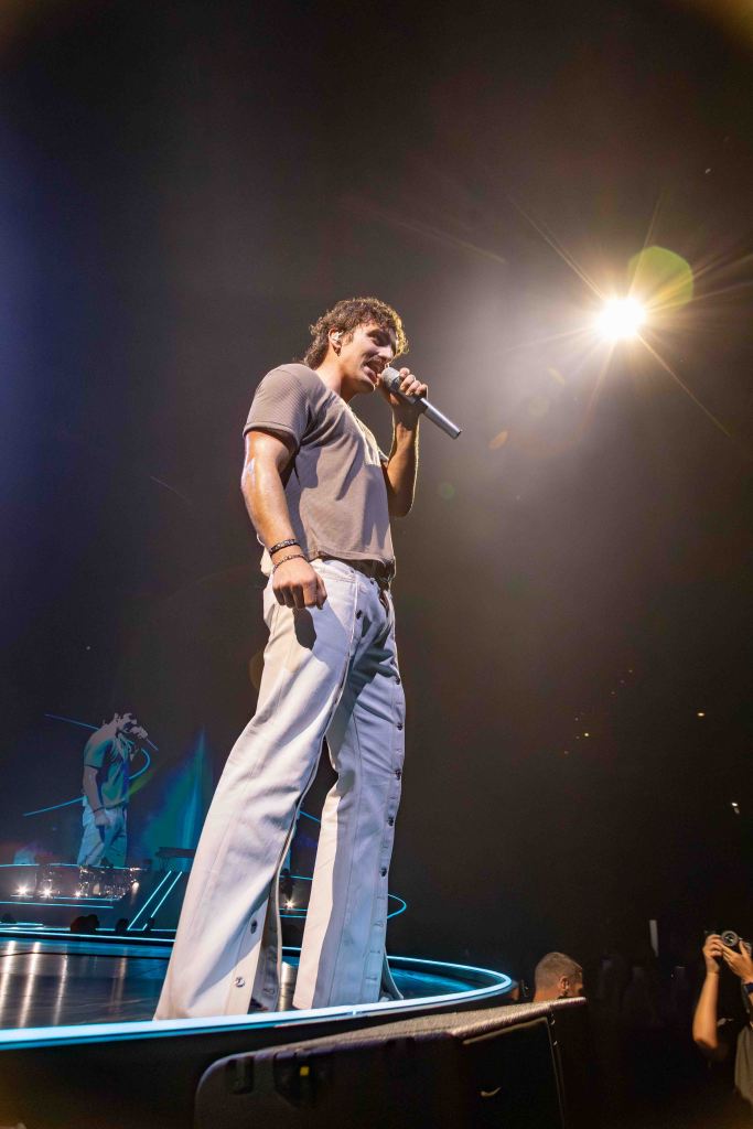 A performer singing on stage, wearing a gray t-shirt and white pants, with bright stage lights and an enthusiastic audience in the background.