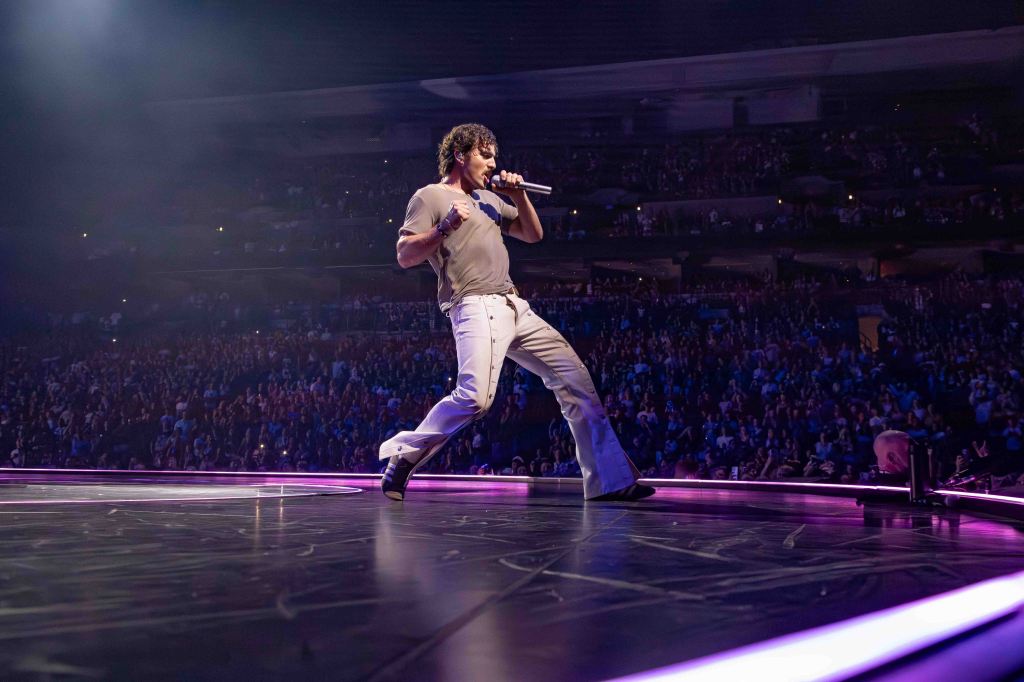 A singer performing on stage with a crowd in the background, holding a microphone and wearing a casual outfit, with dramatic lighting and vibrant colors around.