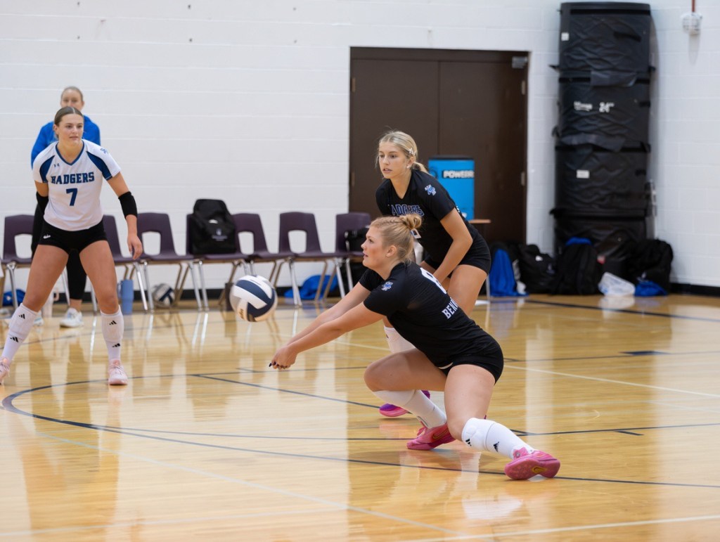 A volleyball player in a black uniform prepares to receive a ball on the court, with two teammates in the background watching the play.