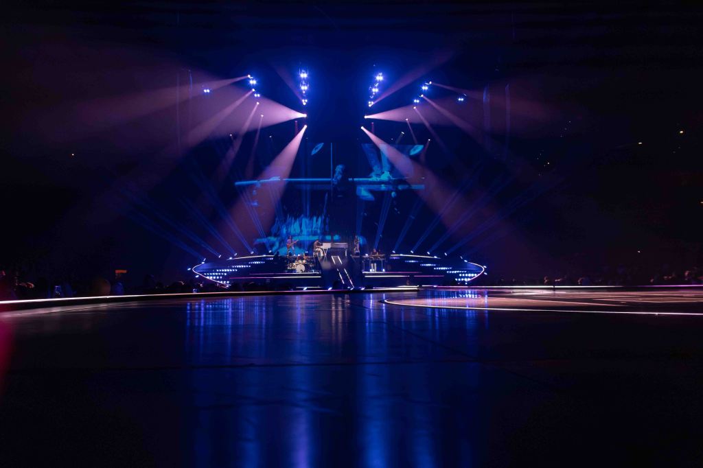 A concert stage illuminated by colorful lights, featuring a circular platform and a band performing in the background.