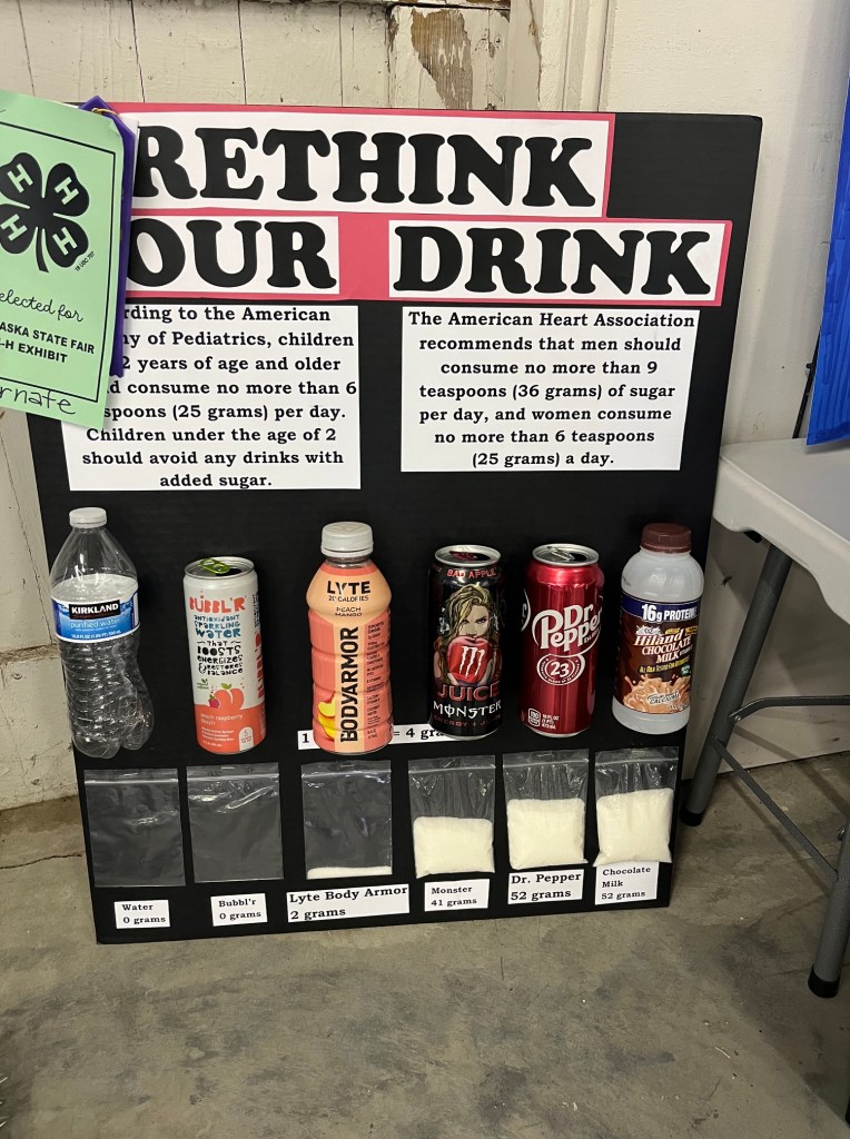 A display board titled 'Rethink Your Drink' featuring various beverages and their sugar content. It highlights recommended daily sugar intake for children and adults, along with images of drinks like water, Lyte Body Armor, Monster, Dr. Pepper, and chocolate milk.
