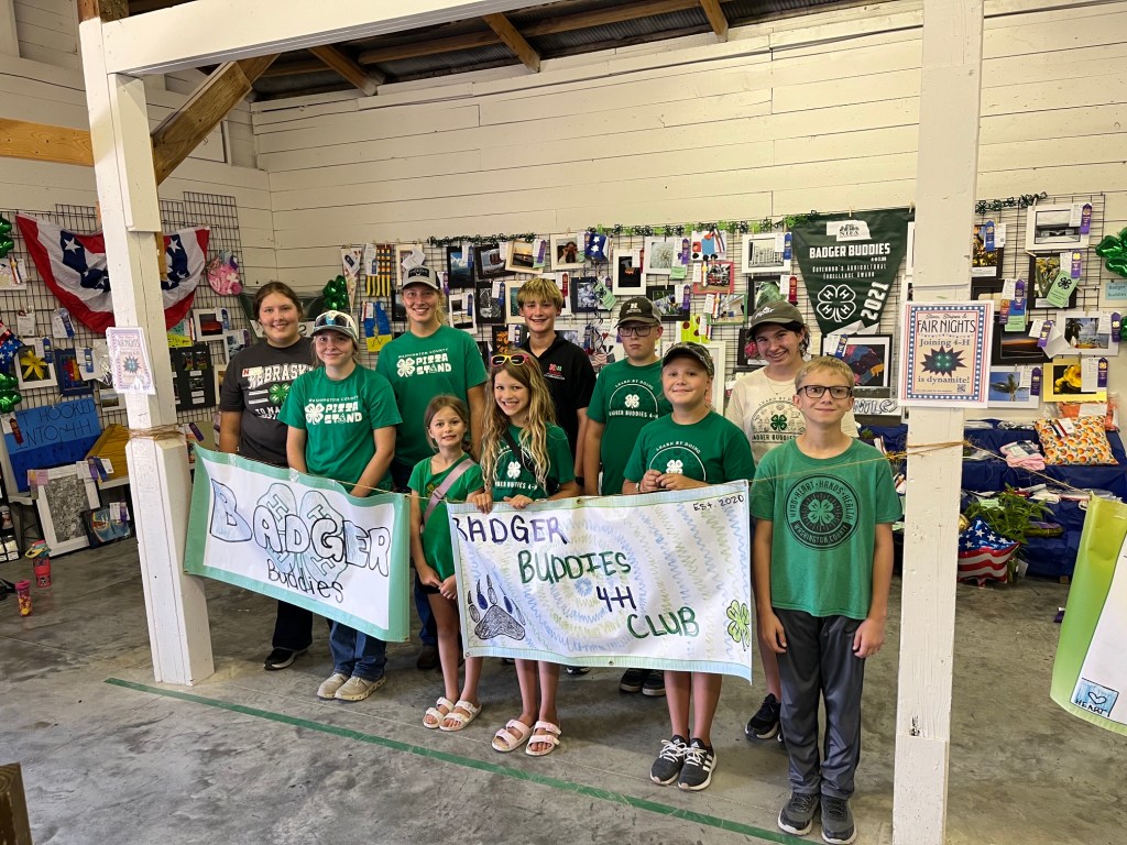 Group of children and adults standing together in a display area, holding banners for 'Badger Buddies' and 'Badger Buddies 4-H Club'. The backdrop features various displayed artworks and ribbons.